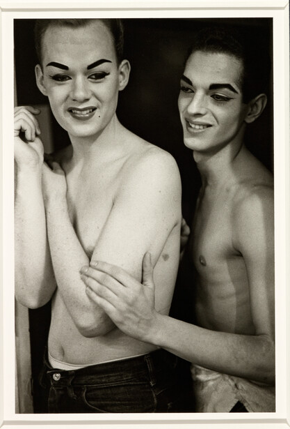 Two shirtless men posing closely together against a dark background, looking at camera with dramatic expressions.