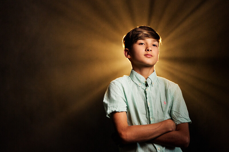 Young boy in light blue button-up shirt looking upward with arms crossed against warm golden background.
