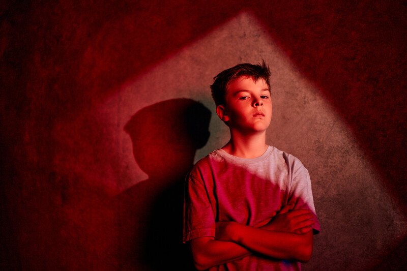 Young person in red shirt standing against red-toned wall with dramatic side lighting and shadow.