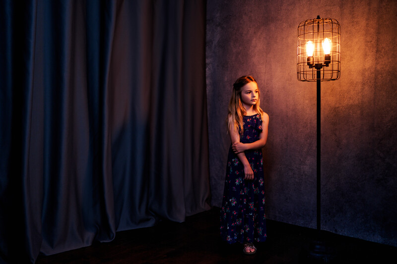 Young woman in dark floral dress standing beside an ornate cage-style floor lamp against textured brown wall with blue curtains.