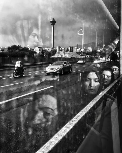 Black and white street scene with rain-covered glass in foreground reflecting faces and city street with cars and tower.