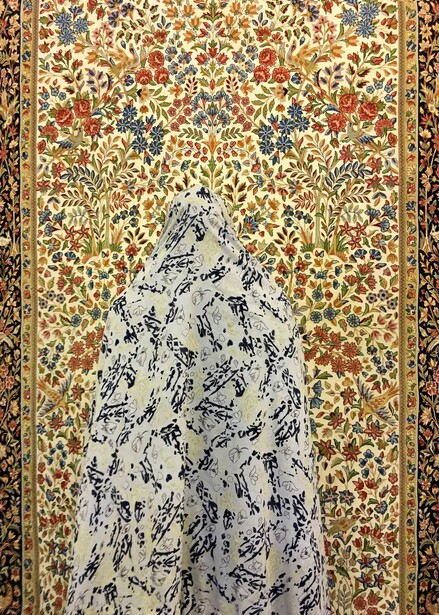 Person in patterned white garment standing against ornate gold and jewel-toned floral textile backdrop.