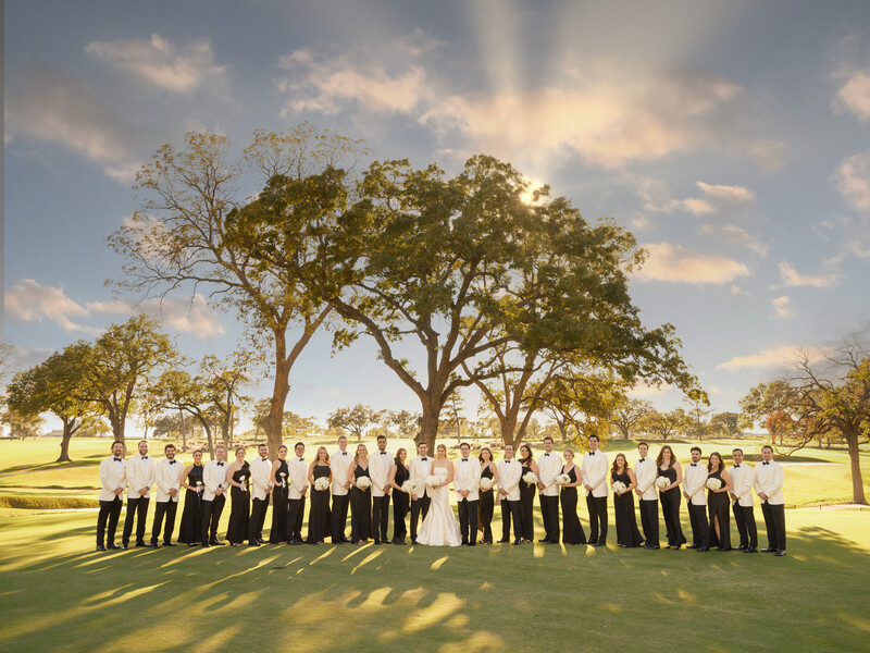 Large group of people in formal black and white attire standing in a line on a sunny grass field with mature trees.