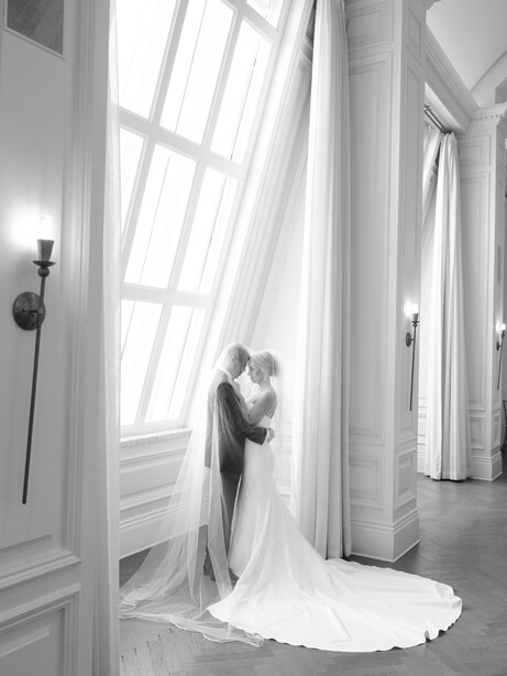 Bride in wedding dress standing at large window with flowing veil in black and white photography.
