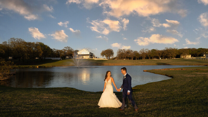 Bride and groom holding hands standing on grass beside a calm lake at sunset.