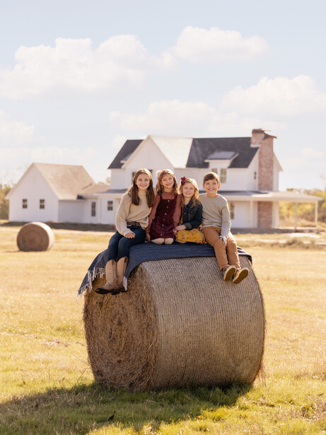 Four people sitting on a large round hay bale in a rural field with farmhouses in the background.