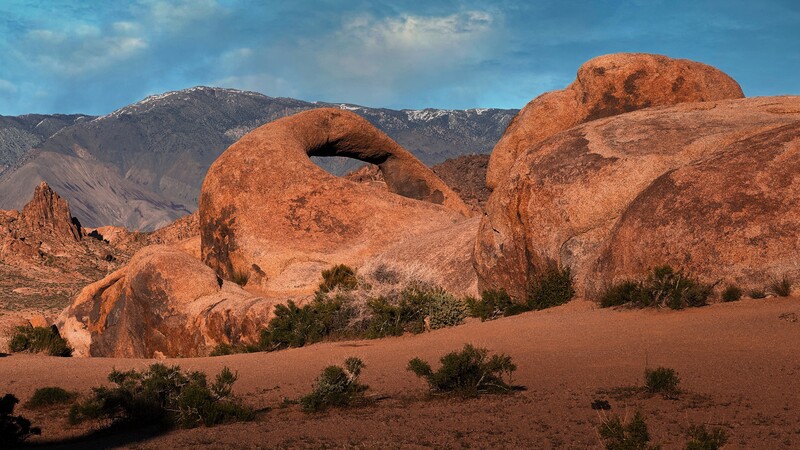 Large rust-colored rock formations with natural arches in a desert landscape beneath snow-capped mountains.