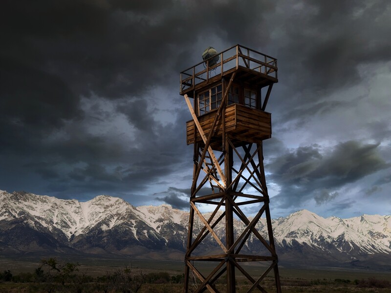 Wooden watchtower structure standing against dramatic storm clouds and snow-capped mountains.