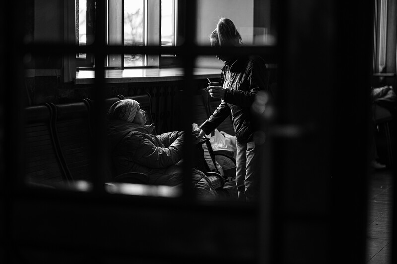 Two people conversing indoors near a window, photographed through a dark foreground element in black and white.