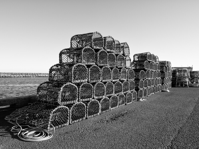 Stacked fishing traps and coiled rope arranged on a pebbled beach in black and white.