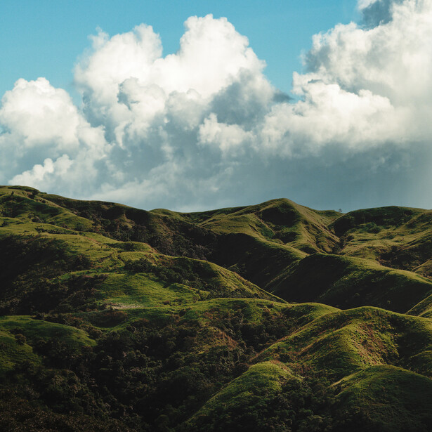 Rolling green hills with dramatic white cumulus clouds under blue sky.