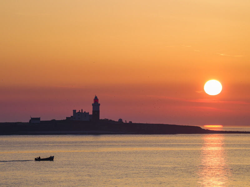 Silhouetted lighthouse on a coastal peninsula at sunset with a small boat in the foreground.