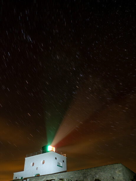 Lighthouse beacon with green and red light beams piercing through a dark night sky filled with stars.