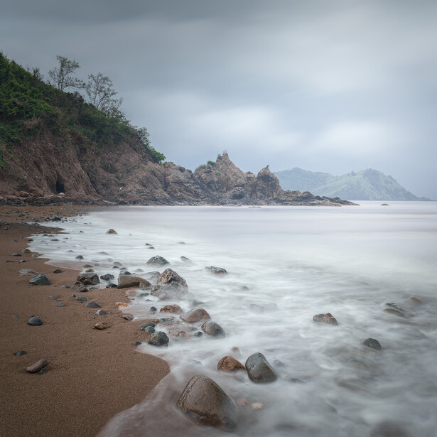 Rocky coastline with long-exposure waves flowing across a sandy beach under overcast sky.