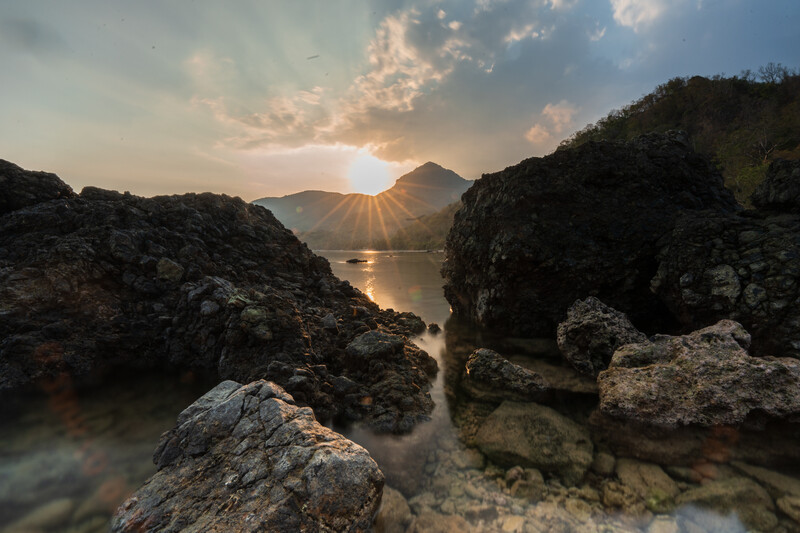 Rocky shoreline with silhouetted boulders framing a bay at sunrise, golden light reflecting on calm water.
