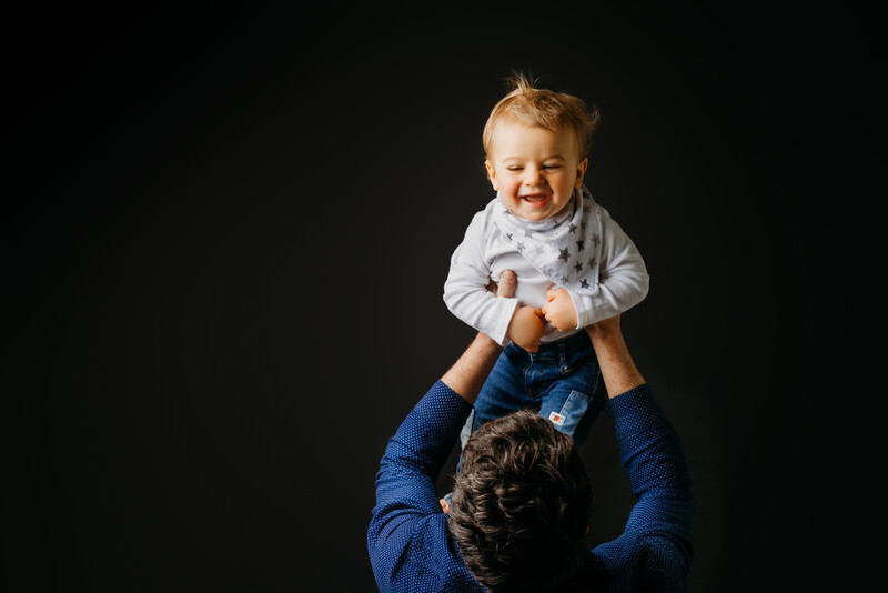 Parent holding a smiling toddler aloft against a black background.