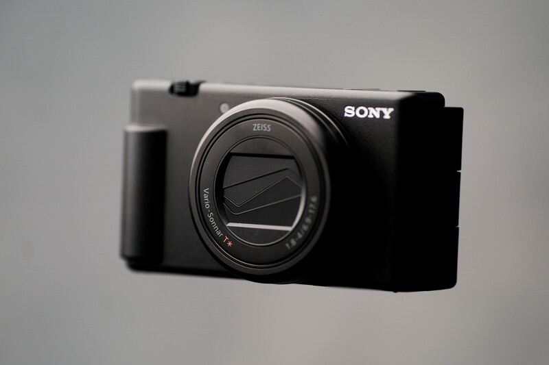 Sony compact camera with Zeiss lens, black body with grip, photographed against neutral gray background.