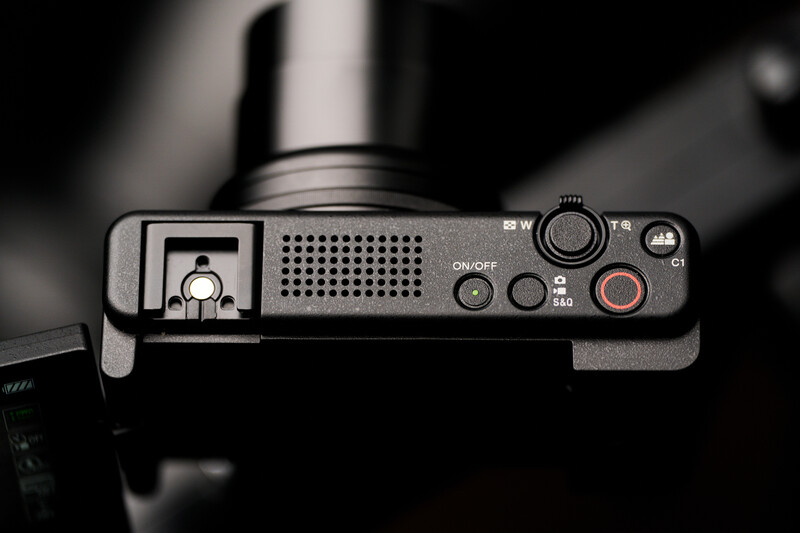Top-down view of a compact camera's control panel showing buttons, dials, and a speaker grille against a dark background.