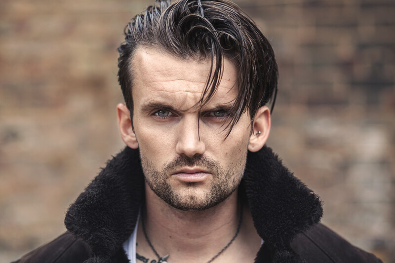 Male portrait with dark swept hair and stubble against blurred brick wall backdrop.