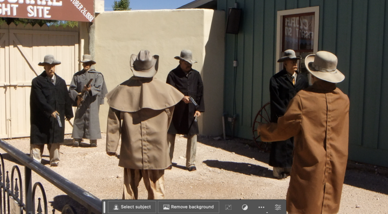 Western-themed scene with costumed actors in period clothing and cowboy hats standing in a dirt courtyard between buildings.
