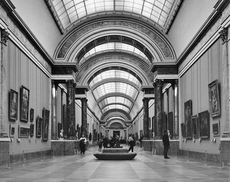 Black and white photograph of a symmetrical gallery corridor with barrel-vaulted skylights and framed artworks lining both walls.
