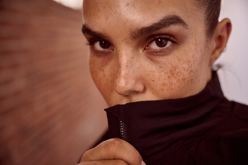 Close-up portrait of a person with freckles and direct gaze, partially obscured by dark fabric pulled up to their face.