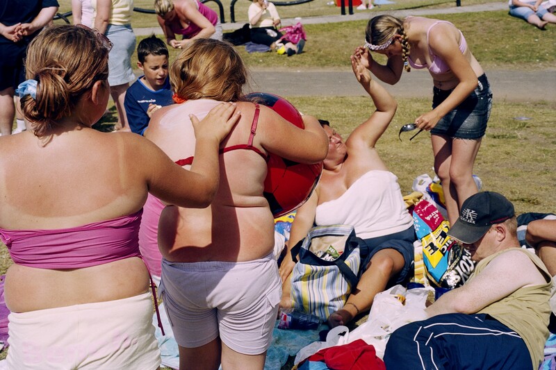 Image shows a family group enjoying fine weather. One user whose skin is red from sun exposure applies sunscreen on another woman's back.