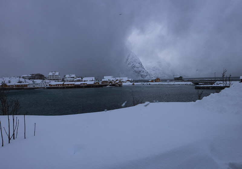 Snow-covered landscape with frozen water and moored ships beneath a dramatic cloudy sky.