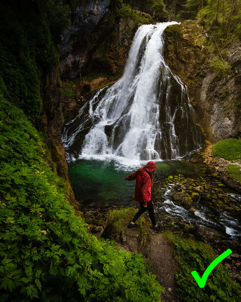 Hiker in red jacket standing before cascading waterfall with emerald pool