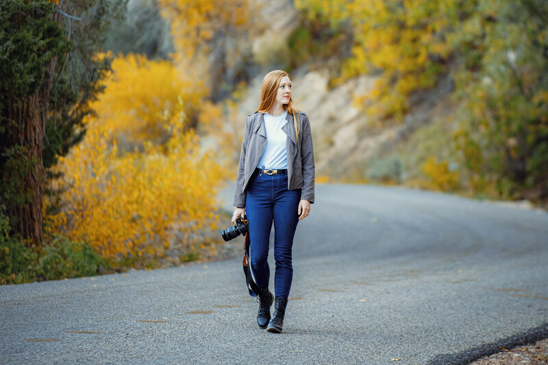 Woman walking along a mountain road lined with golden autumn foliage.