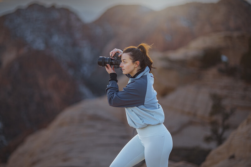 Woman in athletic wear holding a camera to her eye while standing on a rocky overlook.