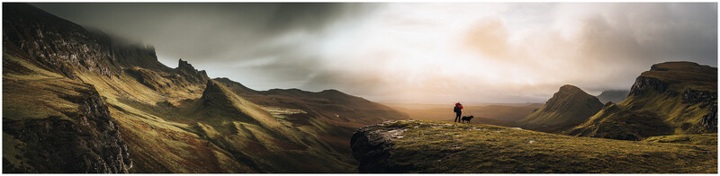 Hiker with dog standing on moss-covered plateau overlooking dramatic mountain valley with layered peaks.