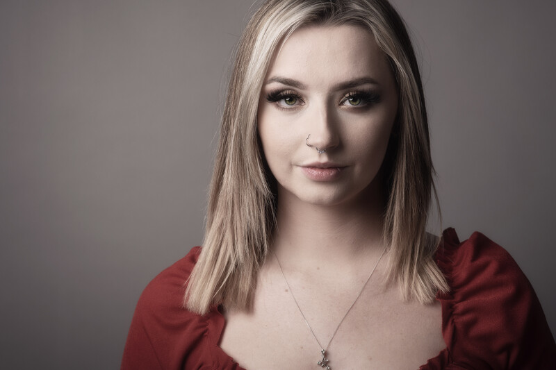 Woman with long blonde hair wearing a red top and pendant necklace against a neutral gray background.