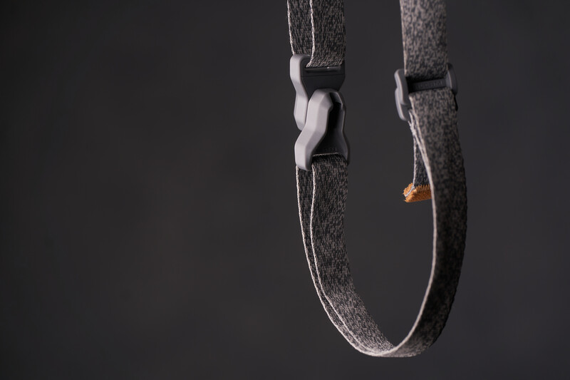Close-up of a gray fabric camera strap with metal hardware and orange accent detail against dark background.