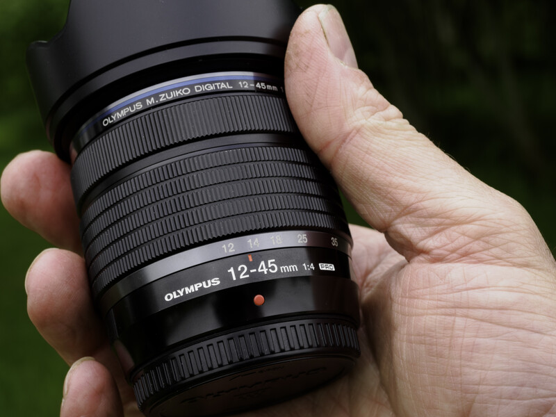Hand holding an Olympus 12-45mm f/3.5-5.6 telephoto zoom lens against a blurred green background.
