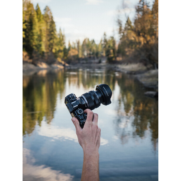 Hand holding a camera with telephoto lens extended toward a forested lake reflection.