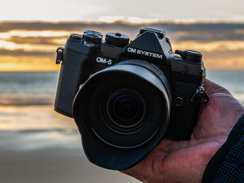 OM-5 camera held in a hand against a beach sunset backdrop.