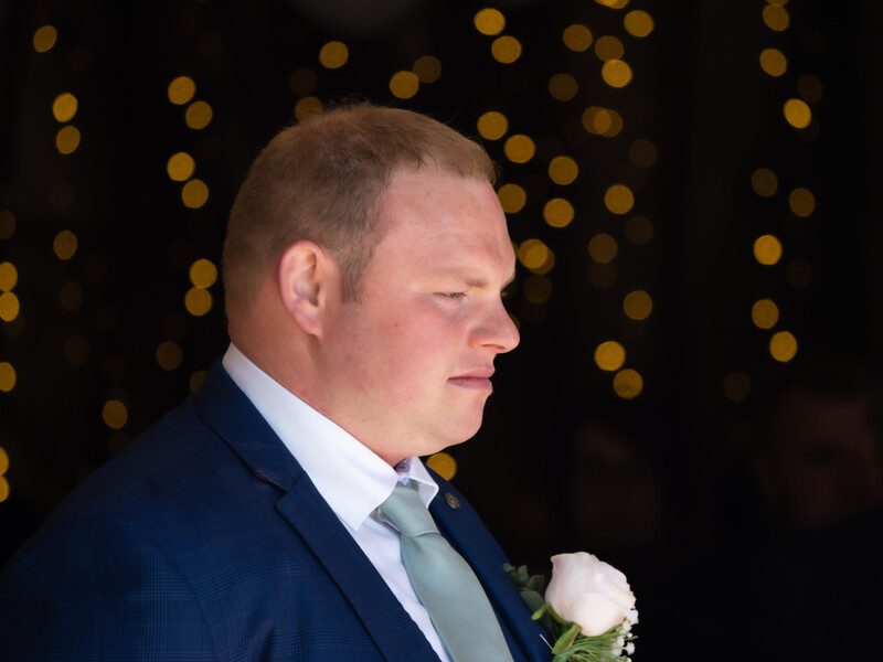 Man in dark suit and light blue tie with white boutonniere, photographed in profile against blurred golden bokeh background.