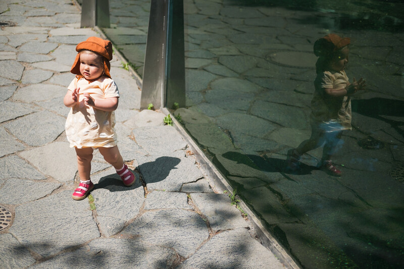 Toddler in orange hat standing on sunlit pavement next to water, with their shadow visible on the water's surface.