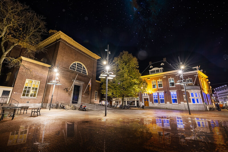 Historic brick buildings illuminated at night in a town square with wet pavement reflecting lights