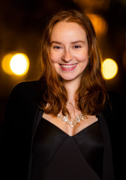 Woman with reddish-brown hair wearing black blazer and statement necklace, photographed against warm golden bokeh background.