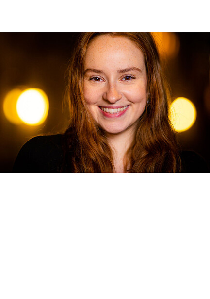 Woman with reddish-brown hair smiling at camera against dark background with warm golden bokeh lights.