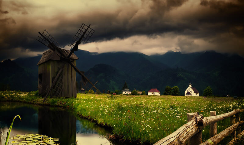 Wooden windmill silhouetted against dramatic storm clouds above a green meadow with scattered houses and mountains in the distance.