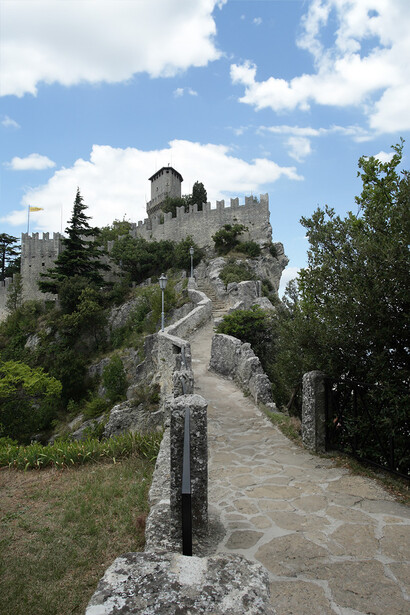 Stone pathway leading uphill toward a medieval fortress with towers and crenellated walls.