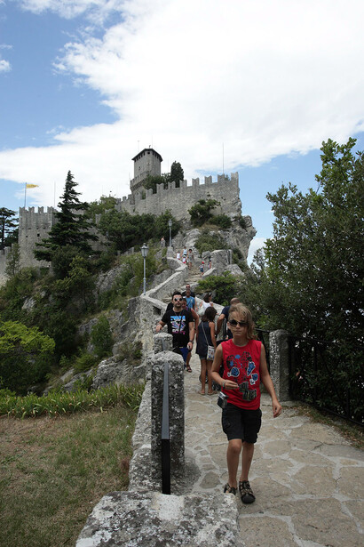 Hikers ascending a stone pathway toward a hilltop fortress with multiple towers under partly cloudy skies.