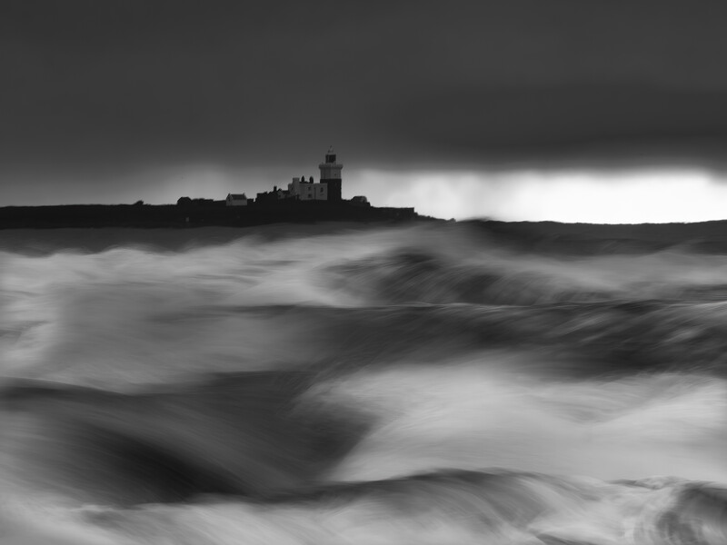 Silhouetted castle on rocky coastline amid turbulent, motion-blurred ocean waves under dramatic storm sky.