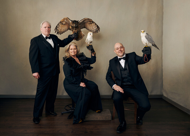 Three people in formal attire posing with falcons against a neutral background.