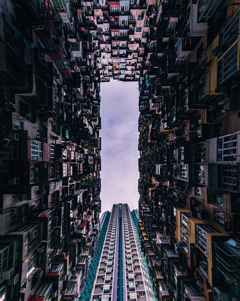 Upward view down a narrow urban street canyon lined with densely packed high-rise buildings framing a sky opening.