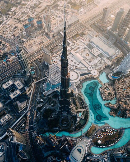 Aerial view of a tall dark skyscraper surrounded by modern buildings and turquoise water features in an urban landscape.