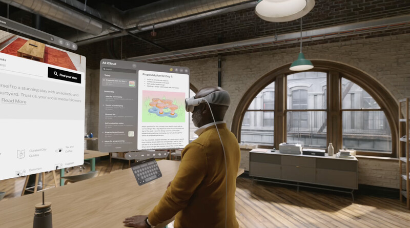 Person in mustard-colored jacket wearing headphones, viewing multiple digital displays in a modern loft workspace.